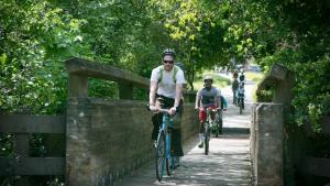 Dos personas sonrientes cruzando un puente peatonal en bicicleta, niños en bicicleta en el fondo