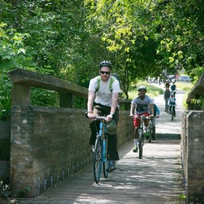 Dos personas sonrientes cruzando un puente peatonal en bicicleta, niños en bicicleta en el fondo