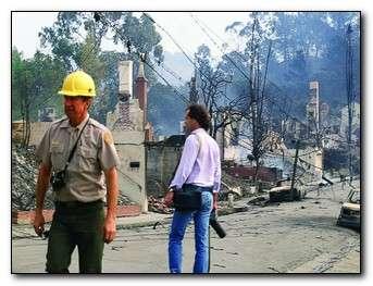 Two people walking around with burned houses in the background