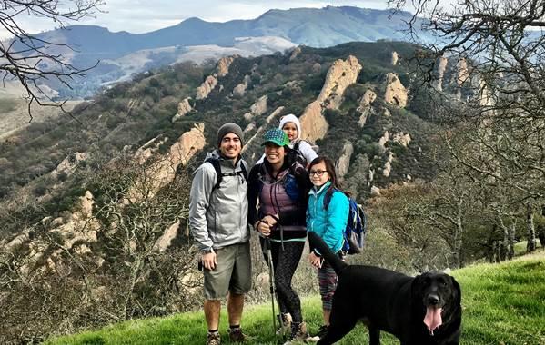 Family of four with dog stands smiling in front of view of rocks and hills